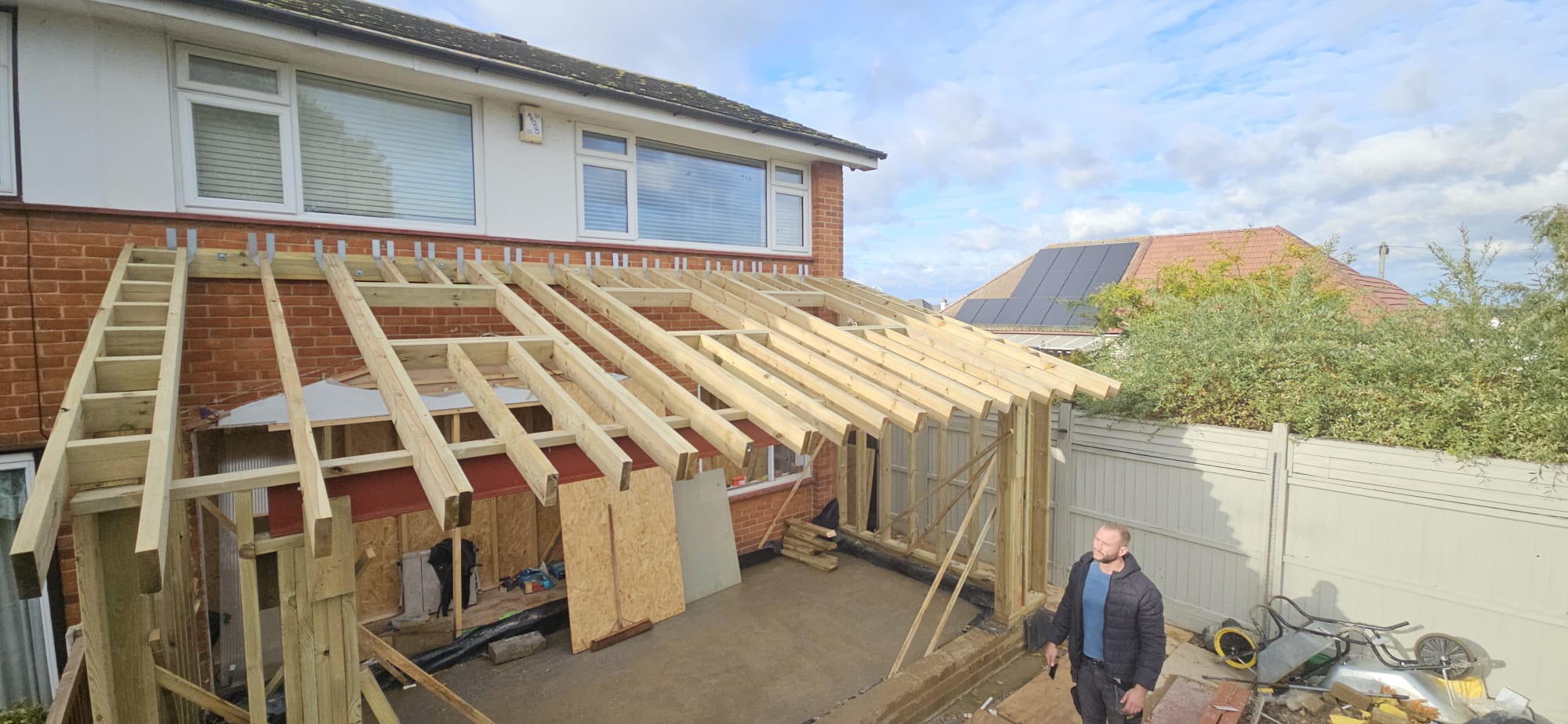 Timber-framed rear extension under construction, with lean-to rafters and roof window openings fixed to a brick and white semi-detached house under a blue sky