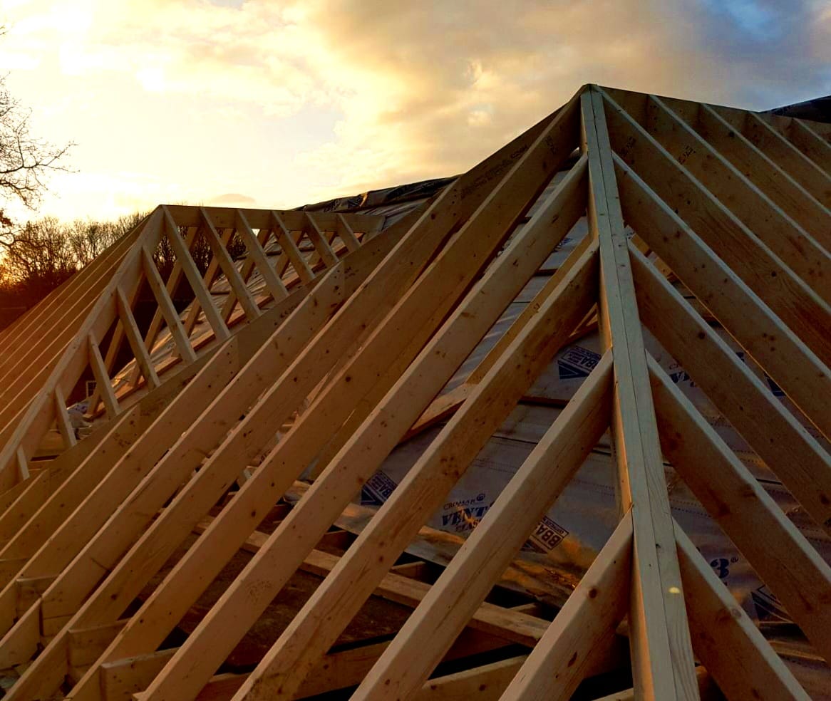 New timber hip roof frame with jack rafters meeting at the hip, photographed from below against a golden sunset sky